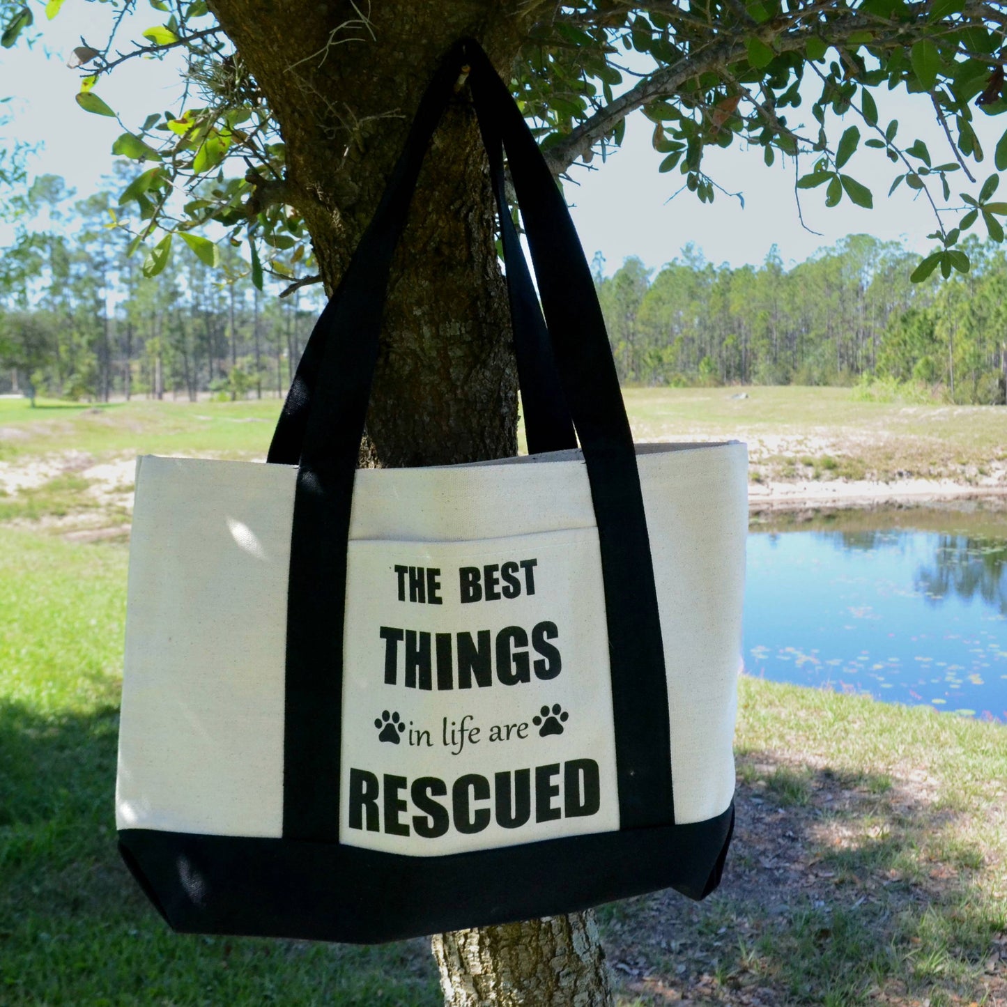 Cotton canvas tote bag. The Best Things in life are Rescued design. Two paw prints, one on each side of the in life are. Handles in black. Background is grass, a small pond, and trees.