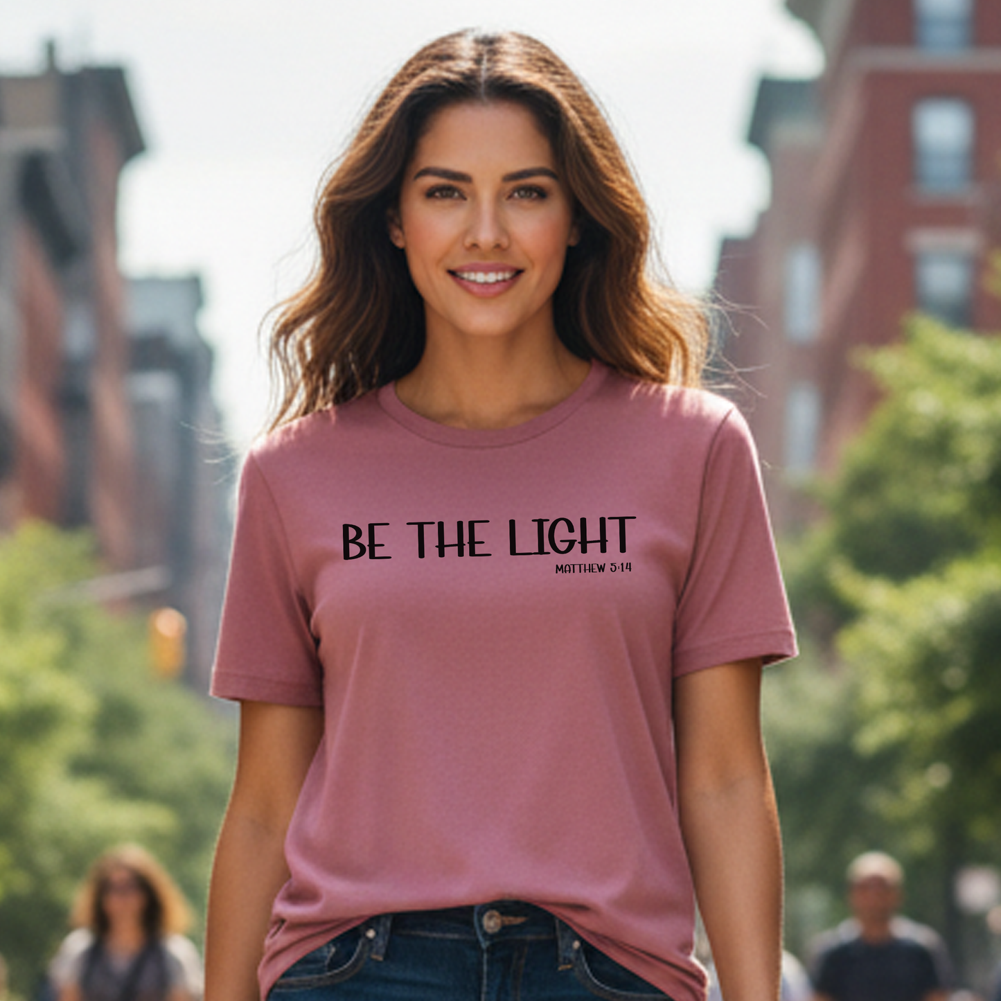 Woman wearing a pink t-shirt with 'BE THE LIGHT' text in an urban setting