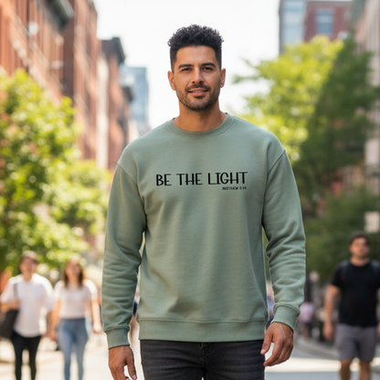 Man wearing a sage color sweatshirt with 'BE THE LIGHT' text on a city street.