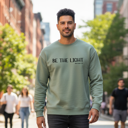 Man wearing a sage color sweatshirt with 'BE THE LIGHT' text on a city street.