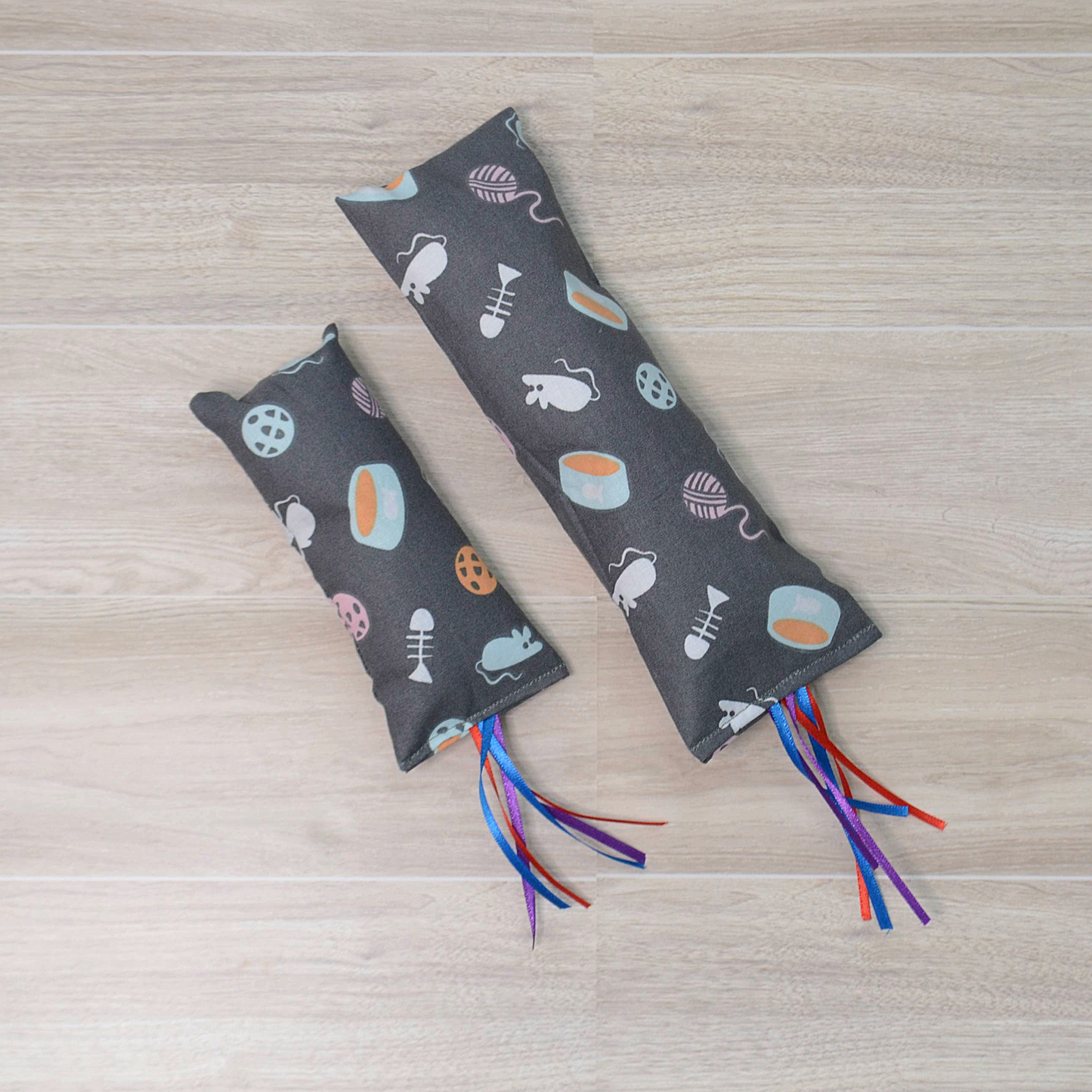Two different size Gray Kitty Kicker toy with mouse and fish  design and colorful ribbon on a wooden surface