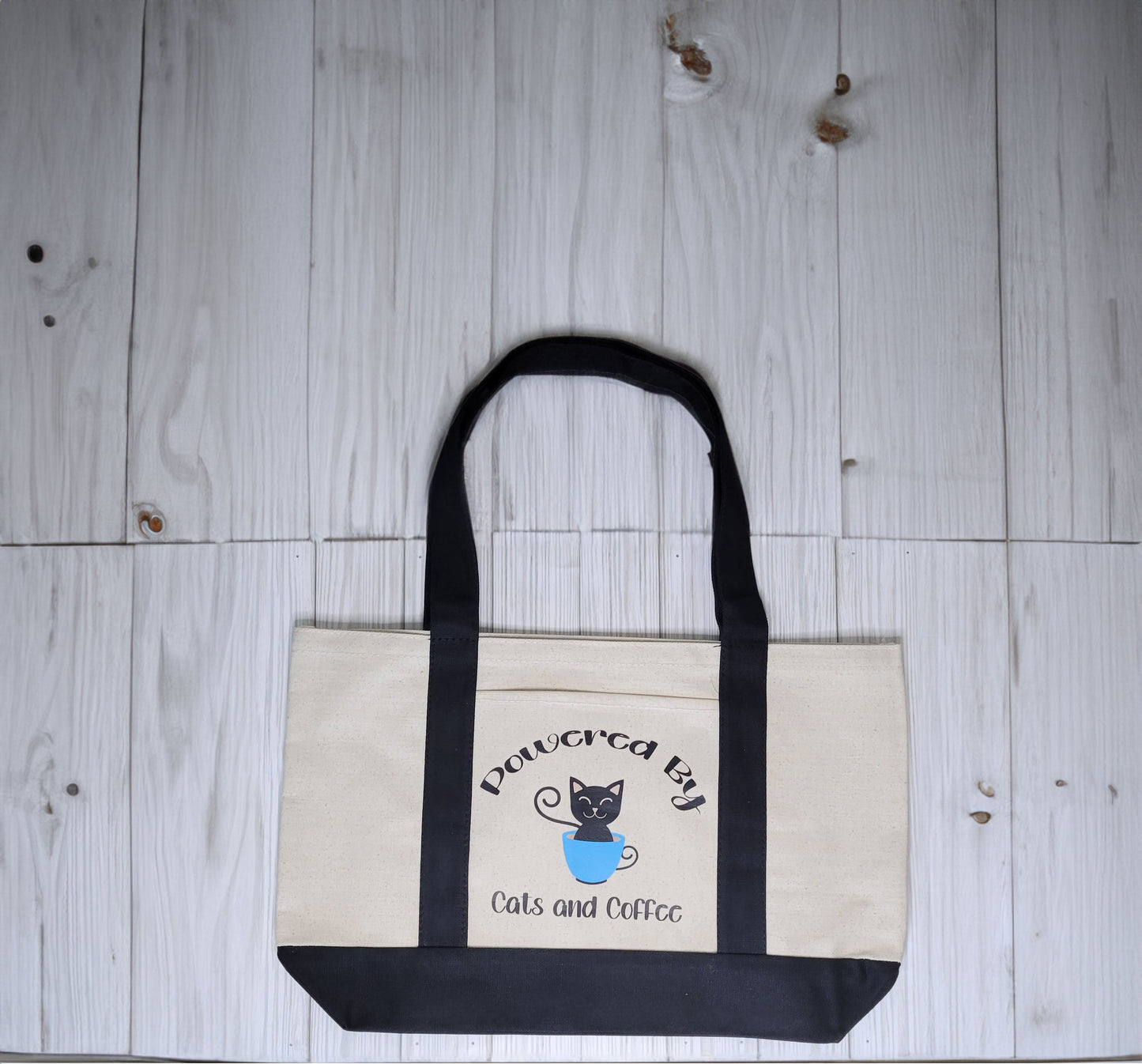 Cotton canvas tote bag with black handles. Includes an outer pocket. Design reads, Powered By Cats and Coffee. Picture of a Black cat inside a blue coffee cup