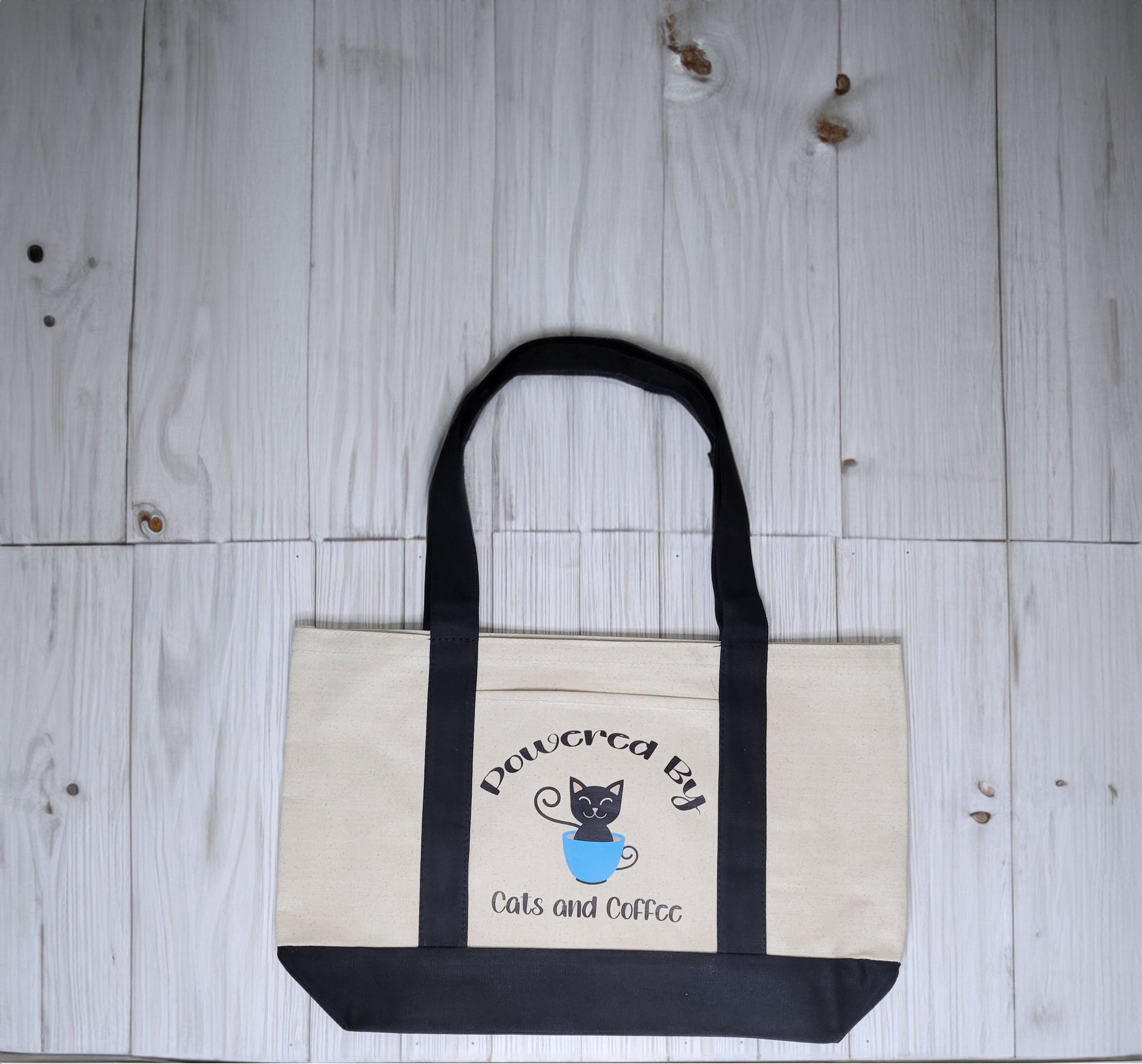 Cotton canvas tote bag with black handles. Includes an outer pocket. Design reads, Powered By Cats and Coffee. Picture of a Black cat inside a blue coffee cup